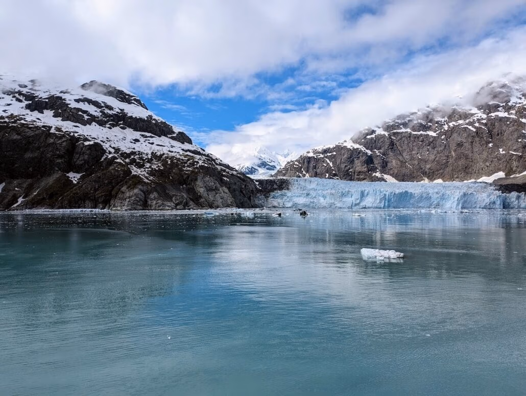 A glacier sits between snow-covered mountains, its blue ice front meeting a calm, reflective body of water. Small ice chunks float beneath a partly cloudy sky—an iconic scene on an Alaska cruise aboard Holland America’s Koningsdam.