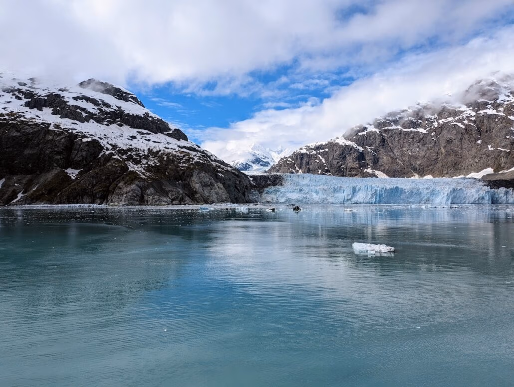 A glacier meets a calm body of water surrounded by snow-capped mountains under a partly cloudy sky. A single piece of ice floats near the center, capturing the serene beauty often seen on an Alaska cruise with Holland America’s Koningsdam.