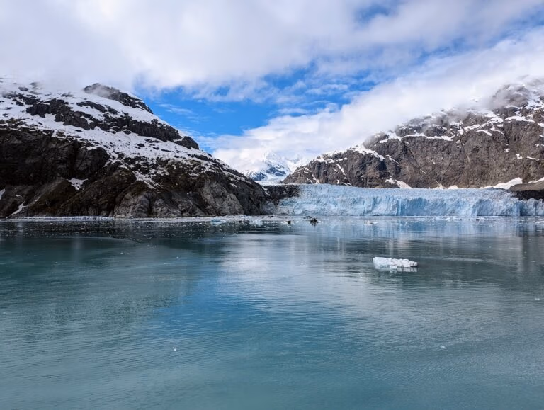 A glacier meets a calm body of water surrounded by snow-capped mountains under a partly cloudy sky. A single piece of ice floats near the center, capturing the serene beauty often seen on an Alaska cruise with Holland America’s Koningsdam.