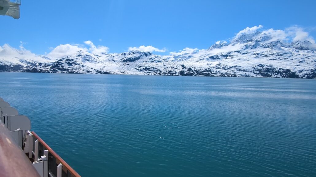 View from Holland America’s Koningsdam, showing calm blue water and a distant snow-covered mountain range under a bright sky. Part of the ship’s railing is visible, capturing the serenity of an Alaska cruise.