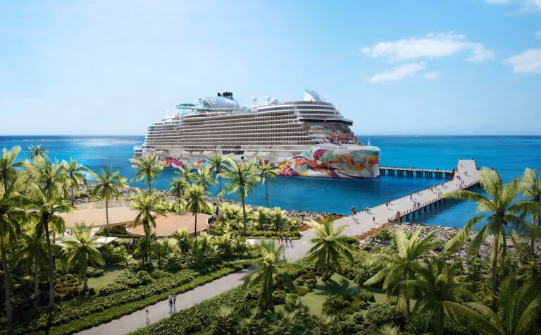 A large cruise ship with colorful artwork on its hull is docked at a pier surrounded by turquoise water, palm trees, and green landscaping under a clear blue sky.