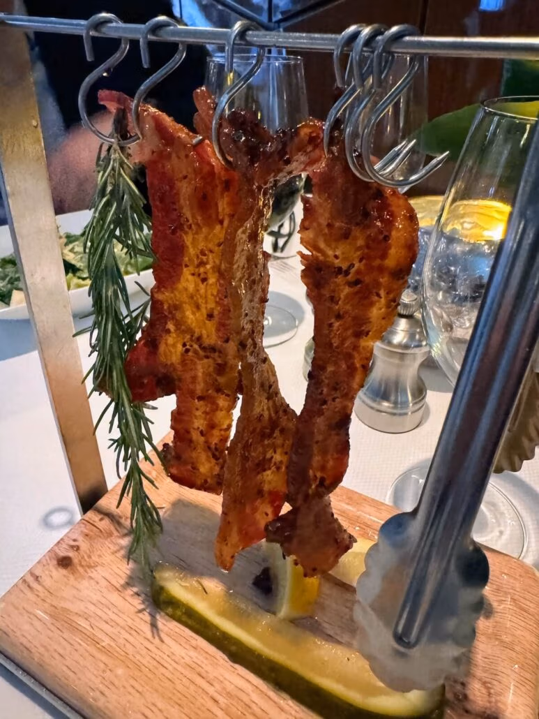 Three strips of cooked bacon hang on metal hooks from a wooden stand, garnished with rosemary and lemon—a savory treat you might find aboard the Koningsdam on your Holland America Alaska cruise, with tableware and glasses in the background.