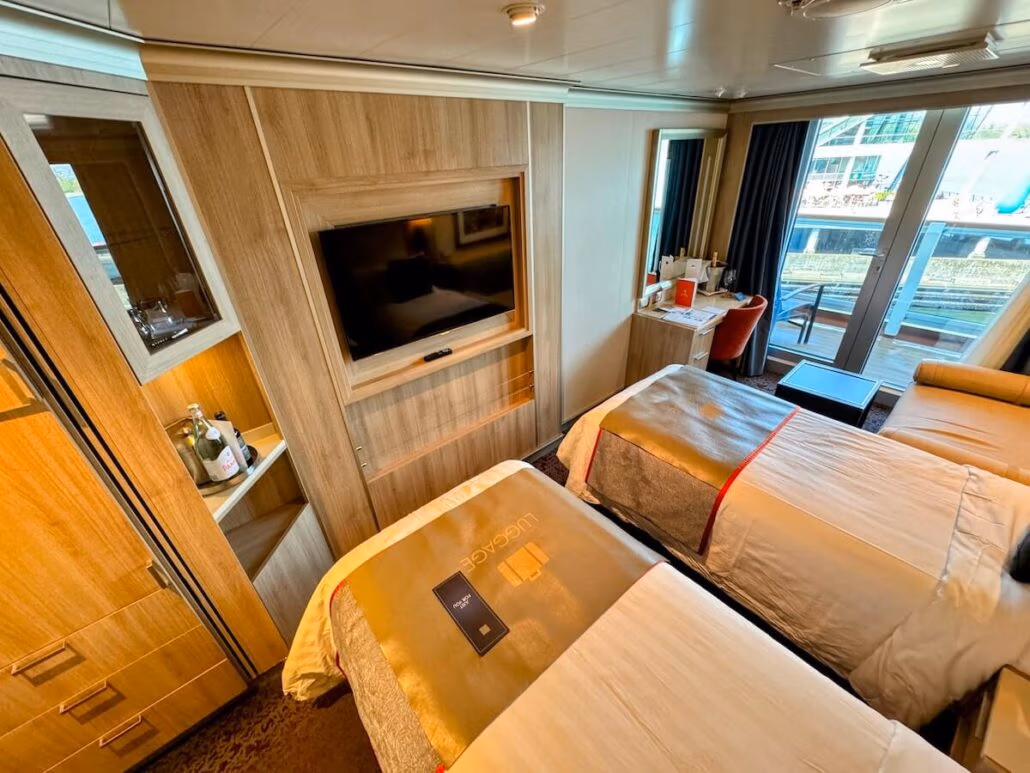 A modern Holland America Koningsdam cruise ship cabin with two single beds, a flat-screen TV, bottled drinks on shelves, a small table near the window, and a sliding glass door opening to your private balcony—perfect for scenic Alaska cruise views.