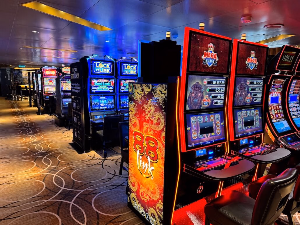 A row of brightly lit slot machines on the Koningsdam casino floor, including red, blue, and black units with digital screens. Patterned carpet and overhead lighting create a lively scene during a Holland America Alaska cruise.