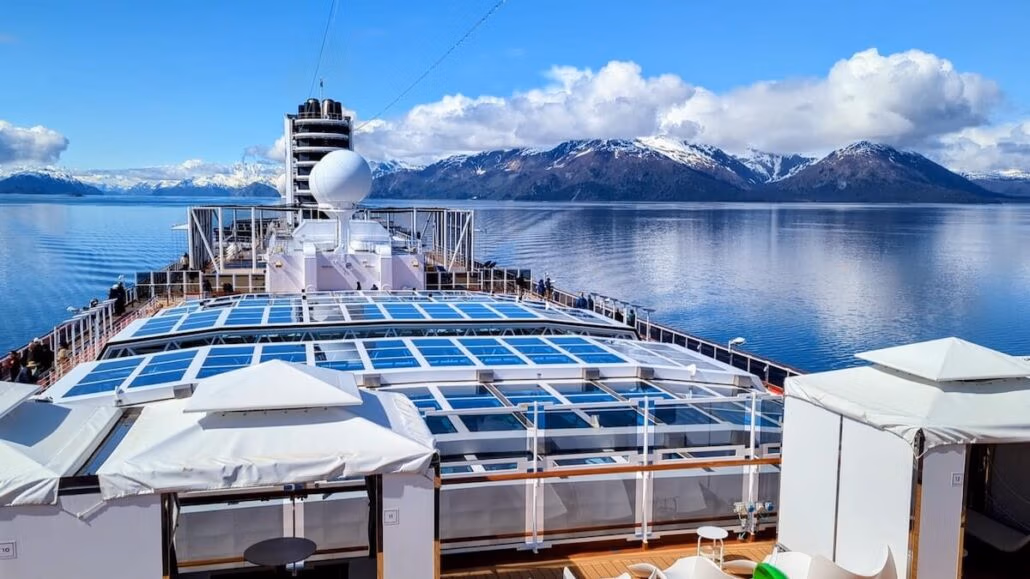 A view from the Holland America Koningsdam cruise ship deck shows calm water and snow-capped mountains in the distance under a partly cloudy sky. The deck features white railings and sunshades, perfect for an unforgettable Alaska cruise.