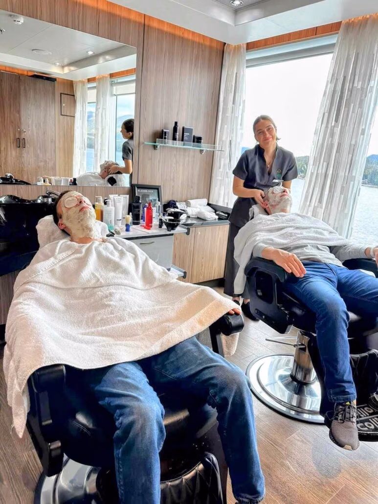 Two people recline in barber chairs with towels and facial masks on their faces, while a person in uniform stands behind them smiling. The modern spa setting, with large windows, could be found aboard the Koningsdam on a relaxing Holland America Alaska cruise.