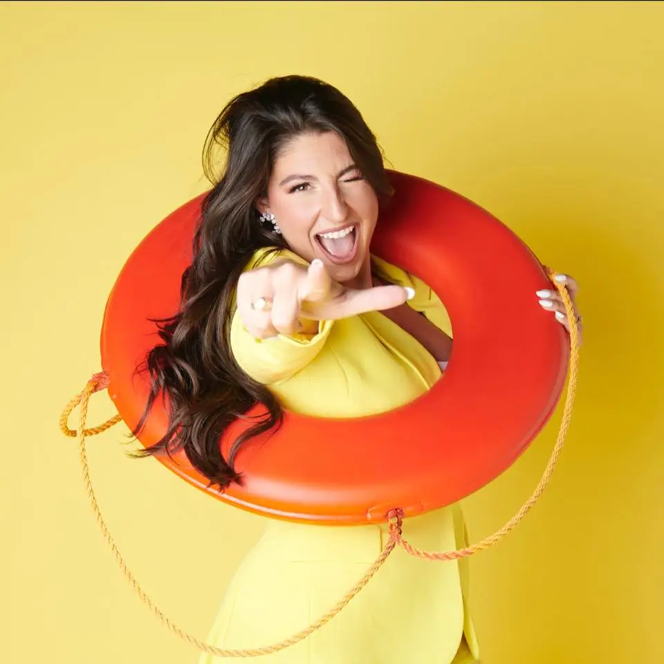 A woman in a yellow blazer stands against a yellow background, smiling and winking while pointing at the camera. Posing as a Carnival Cruise director, she has a red life preserver ring around her shoulders.