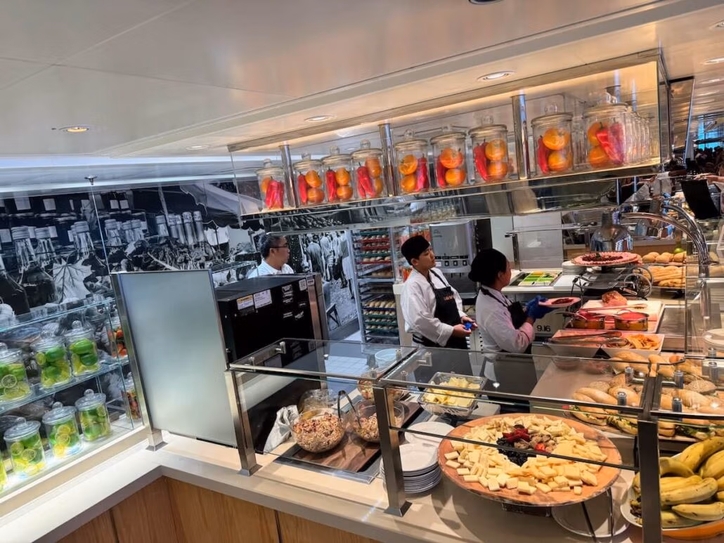 A buffet counter aboard Koningsdam features two staff behind glass displays, offering cheese, fruit, pastries, and jars of preserves. A black-and-white mural on the wall adds charm to this Holland America Alaska cruise dining experience.