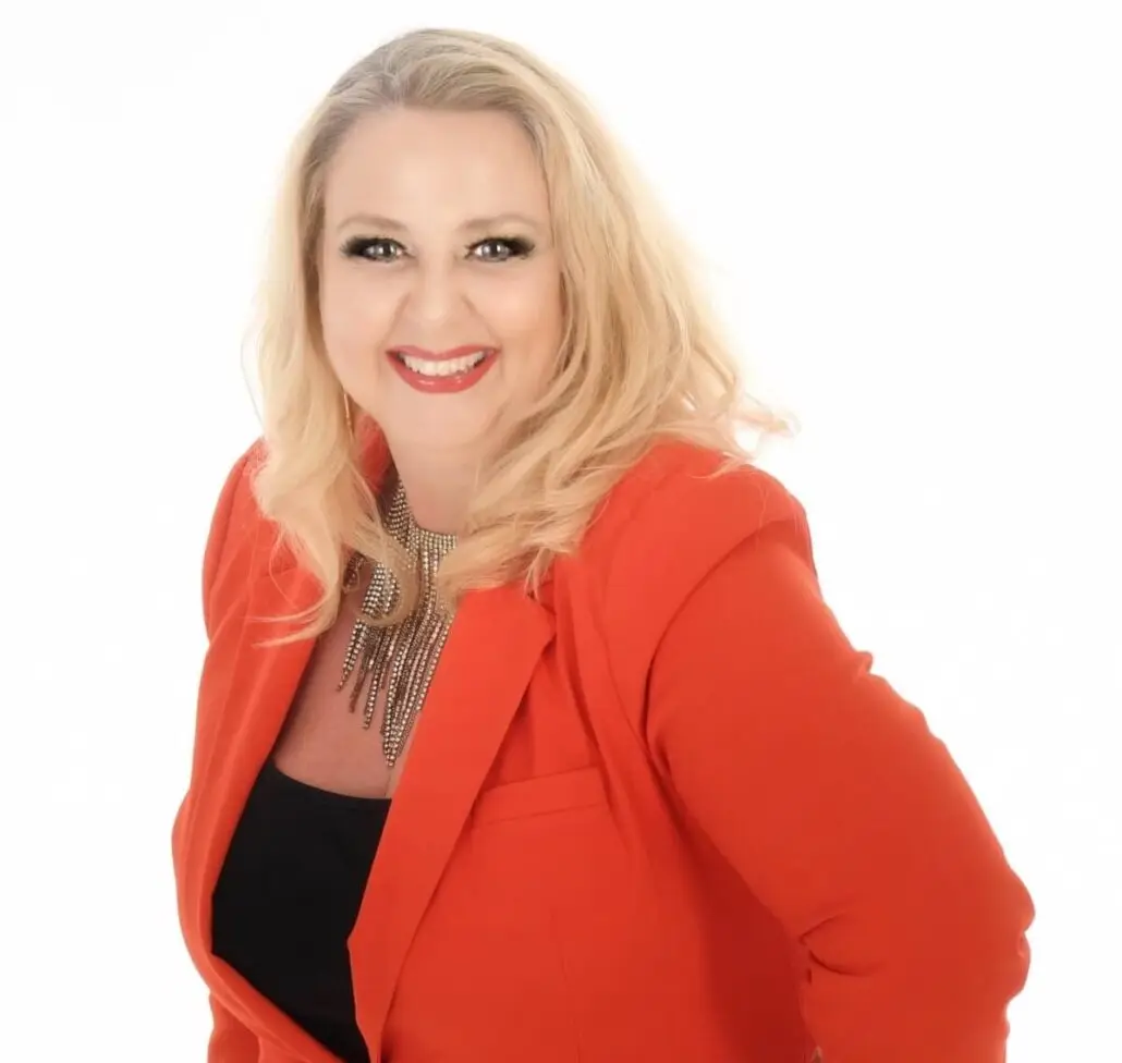 A woman with long blonde hair, dressed in a bright red blazer over a black top and statement necklace, smiles and poses against a white background—resembling the confident style of a Carnival Cruise Director.