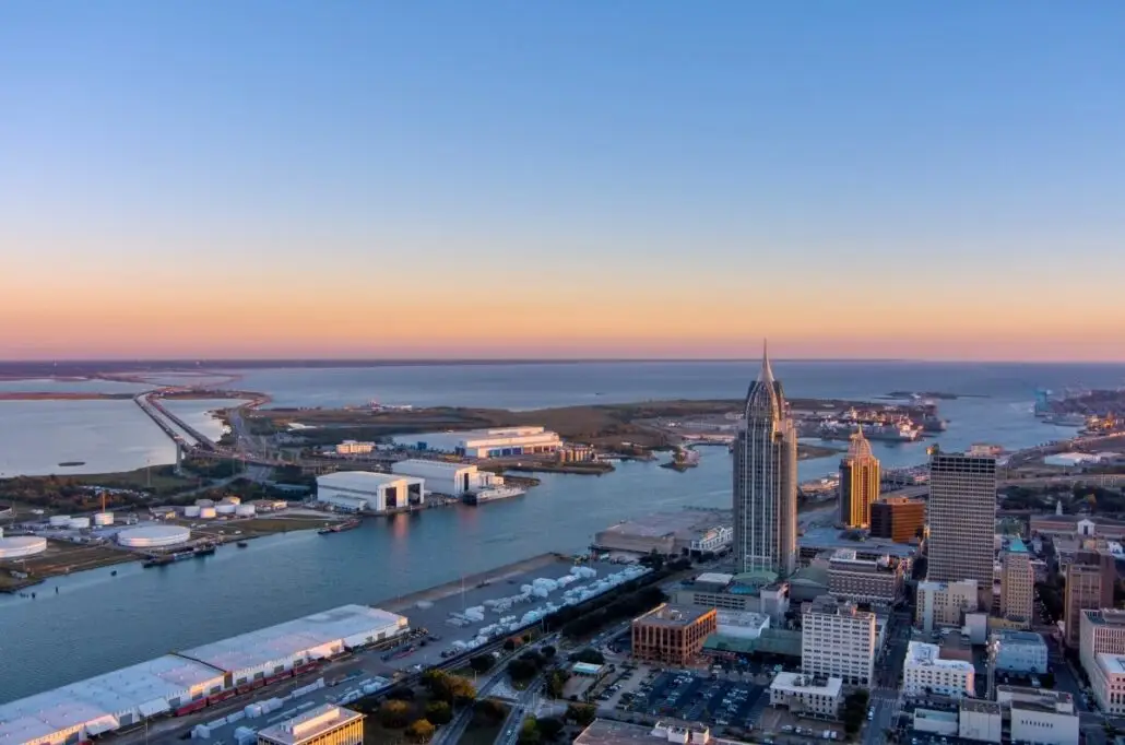 Aerial view of downtown Mobile, Alabama, at sunset, showing tall buildings by the waterfront, industrial areas, bridges over the bay, and the bustling port where Mobile Alabama cruises will set sail in 2026.