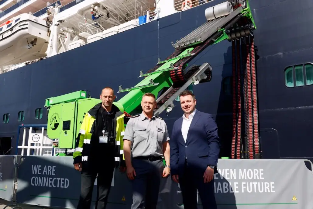 Three men stand in front of Holland America’s Nieuw Statendam near a green piece of shore power machinery. Two banners on the railing read, “WE ARE CONNECTED” and “EVEN MORE VIABLE FUTURE.” The group is outdoors in daylight.