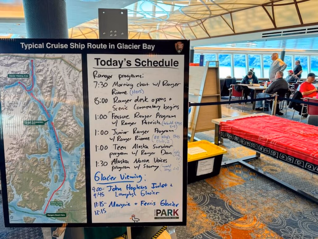 A whiteboard displays a Glacier Bay Alaska cruise route map and a schedule of ranger programs and activities. In the background, people sit at tables with a red patterned tablecloth, perfect for Holland America Koningsdam guests exploring Alaska.