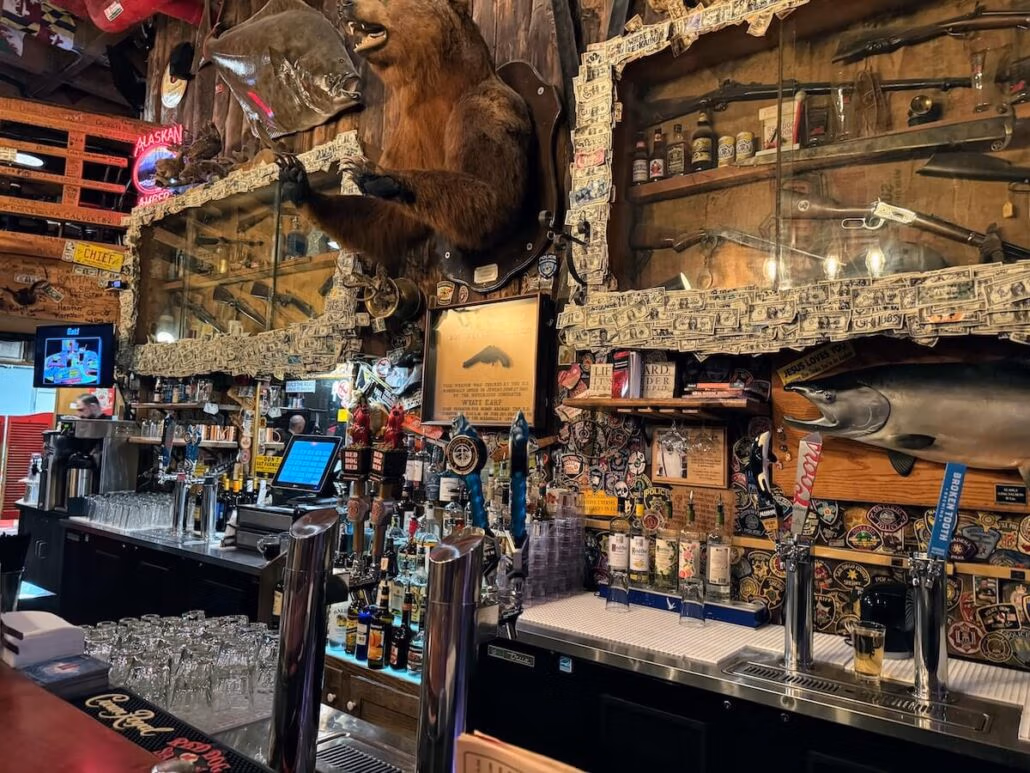 A bar aboard the Koningsdam with two mirrors framed in dollar bills, a bear taxidermy above the counter, various bottles on shelves, beer taps, a swordfish mount, and eclectic decorations—a true Holland America Alaska cruise experience.
