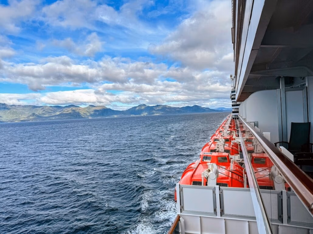 A Holland America cruise ship sails across the ocean, its rows of red lifeboats visible as distant mountains and clouds frame the partly cloudy sky—an unforgettable Alaska cruise scene.
