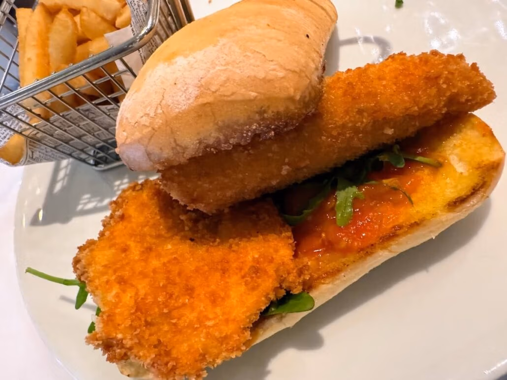 A sandwich with breaded and fried fish fillets, green leafy vegetables, and sauce on a bun, served on a white plate with French fries in a small metal basket—a tasty bite inspired by Holland America’s Alaska cruise aboard Koningsdam.