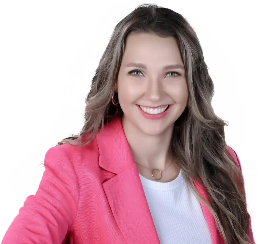 A woman with long wavy brown hair, wearing a pink blazer over a white top, smiles at the camera—looking ready to announce the 2025 Carnival Cruise Director Schedule against a plain white background.