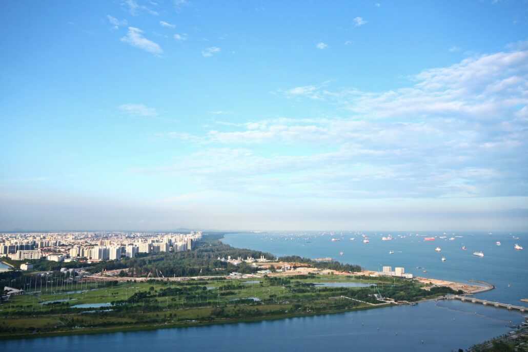 A coastal cityscape featuring a mix of tall buildings, green spaces, and a shoreline with many ships anchored offshore under a blue sky with scattered clouds.