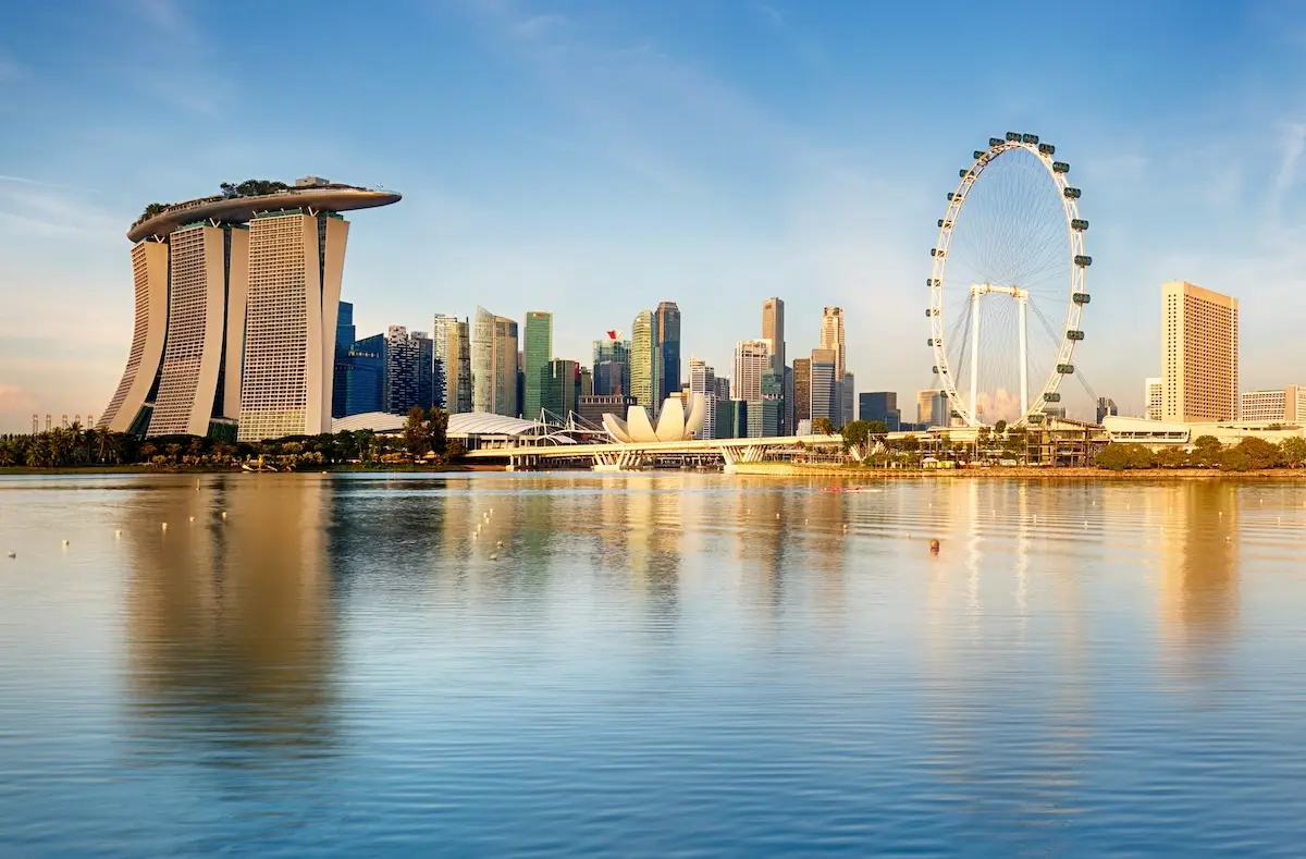 Skyline of Singapore with Marina Bay Sands, the Singapore Flyer Ferris wheel, and modern high-rise buildings reflecting in calm water under a clear blue sky.