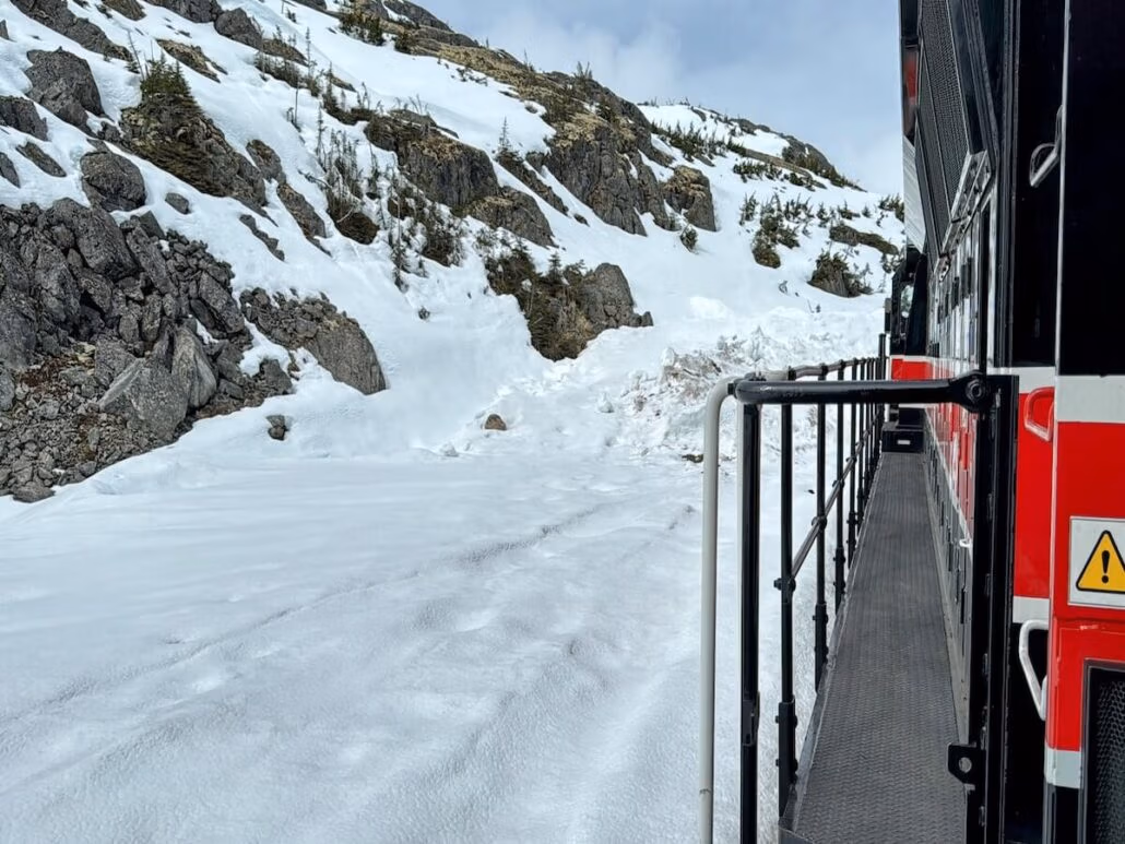 A red and black train travels through a snowy, mountainous Alaskan landscape with rocky slopes and scattered trees—an unforgettable scene often glimpsed on a Holland America Alaska cruise excursion, with the view captured from the side of the train and its metal railing.