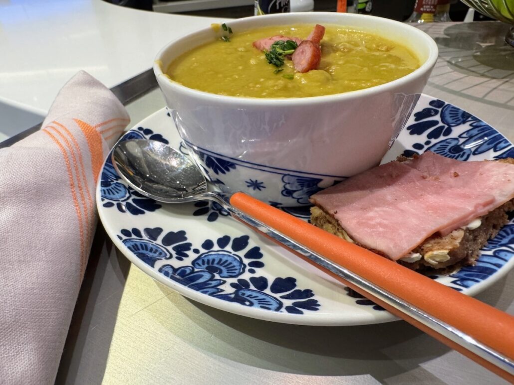 A blue and white plate holds a bowl of yellow soup with sausage and herbs, a slice of bread with ham, an orange-handled spoon, and a folded napkin—capturing the refined dining experience aboard a Holland America Alaska cruise.