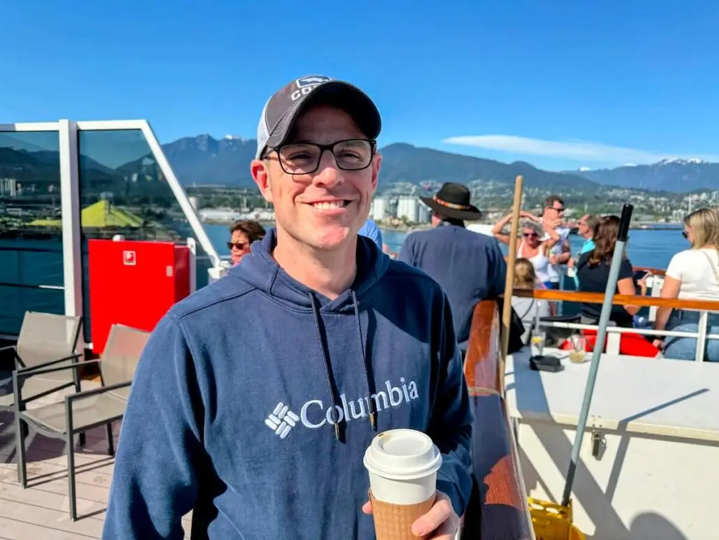 A man in a blue Columbia hoodie, glasses, and a baseball cap smiles with a coffee cup on the sunny deck of Koningsdam during a Holland America Alaska cruise, with mountains, a city, and other passengers in the background.