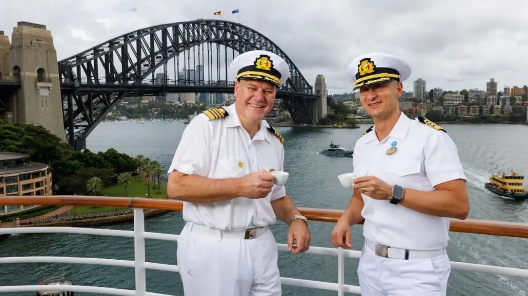 Two cruise ship captains in white uniforms and caps stand on a deck, holding coffee cups, with the Sydney Harbour Bridge and water in the background on a cloudy day, sharing stories of Asia sailing adventures.
