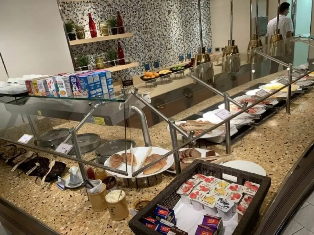 A Carnival breakfast buffet features boxed cereal, yogurt cups, butter, bread, hot dishes under heat lamps, and fresh fruit on a counter, with bowls, plates, and serving utensils arranged neatly. Please note any relevant food warning labels displayed.