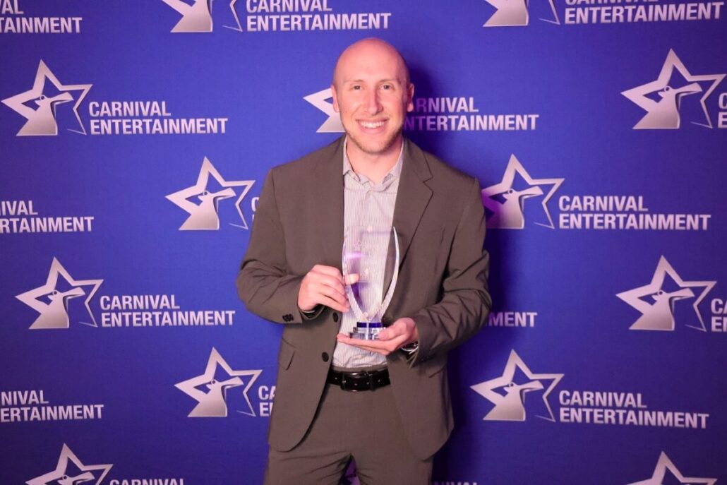 A fan-favorite cruise director in a brown suit stands before a blue Carnival Entertainment banner, holding a clear trophy and smiling at the camera.