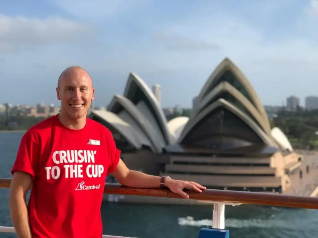 A fan-favorite cruise director wearing a red “Cruisin’ to the Cup” T-shirt stands on a railing with the Sydney Opera House in the background on a sunny day.