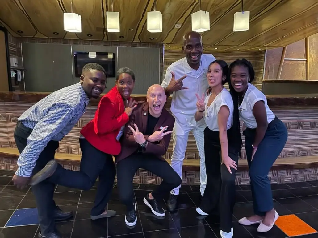 Six people pose enthusiastically in front of a wooden bar, smiling and making peace signs. Dressed in business or business-casual attire, they channel the energy of a Carnival fan-favorite cruise director for a fun, memorable moment.