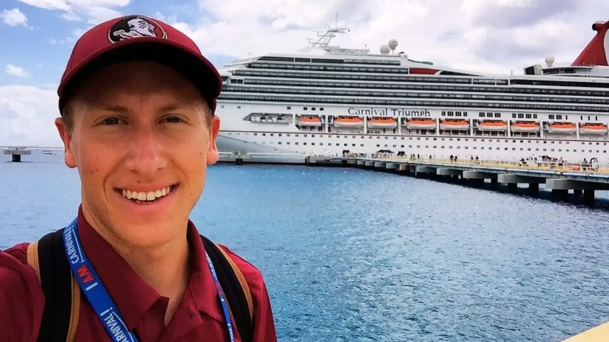 A man wearing a maroon cap and shirt smiles at the camera with the Carnival cruise ship "Carnival Triumph" docked behind him over blue water under a partly cloudy sky, capturing a Fan-Favorite moment.
