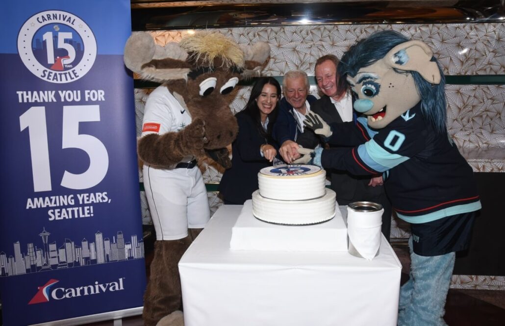 Four people, including two mascots in costume, stand around a cake and cut it together. A banner next to them reads "Carnival 15 Seattle: Thank you for 15 Amazing Years, Seattle!"—celebrating Carnival Luminosa’s Alaska cruise season.