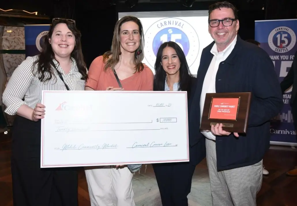 Four people smile indoors as two women hold a $20,000 check from Carnival Cruise Line, celebrating a Carnival Luminosa Alaska cruise in Seattle. A man displays a framed plaque, with the Carnival Cruise Line logo visible behind them.