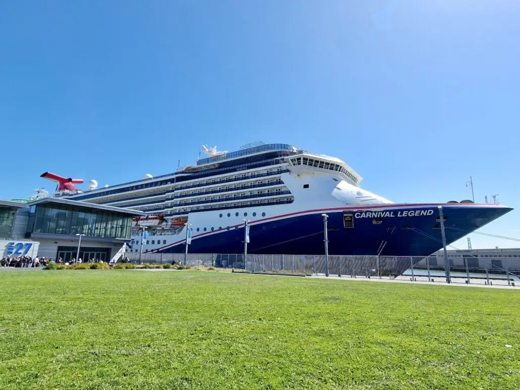 A large cruise ship named Carnival Legend is docked at a port terminal in San Francisco next to a grassy area under a clear blue sky.