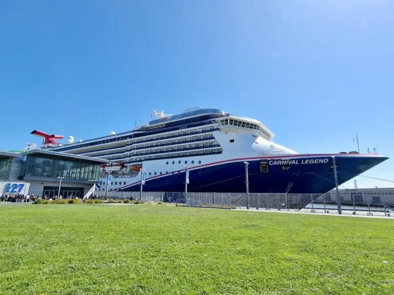 A large cruise ship named Carnival Legend is docked at a port terminal in San Francisco next to a grassy area under a clear blue sky.