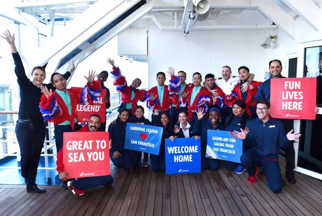 A group of people on a cruise ship pose and smile, holding colorful signs that say "CARNIVAL LEGEND," "GREAT TO SEA YOU," "WELCOME HOME," "FUN LIVES HERE," and others with celebratory text.