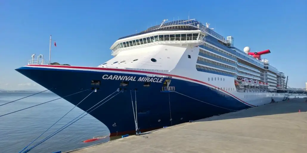 The Carnival Miracle, featuring new features from its recent dry dock, is a large cruise ship docked at the port. Painted dark blue and white with red accents, it stands out against the clear blue sky above.