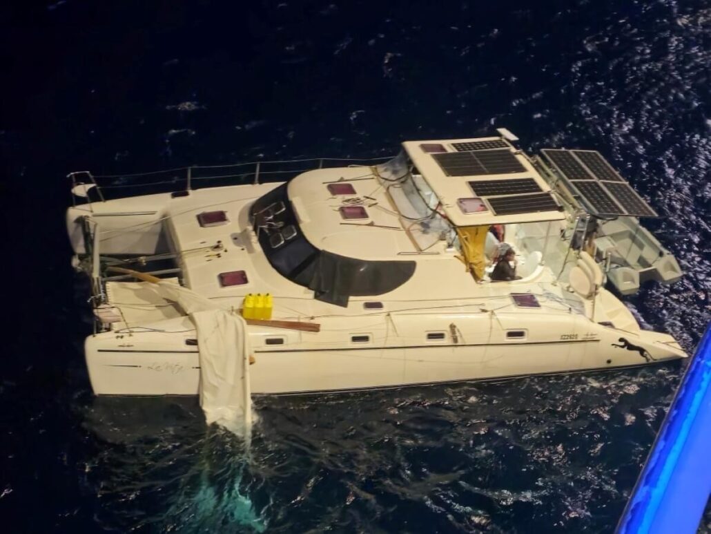 A stranded catamaran with solar panels and yellow jerry cans floats in dark blue water at night. A man sits near the helm while a damaged sail hangs over the side, awaiting cruise ship rescue.