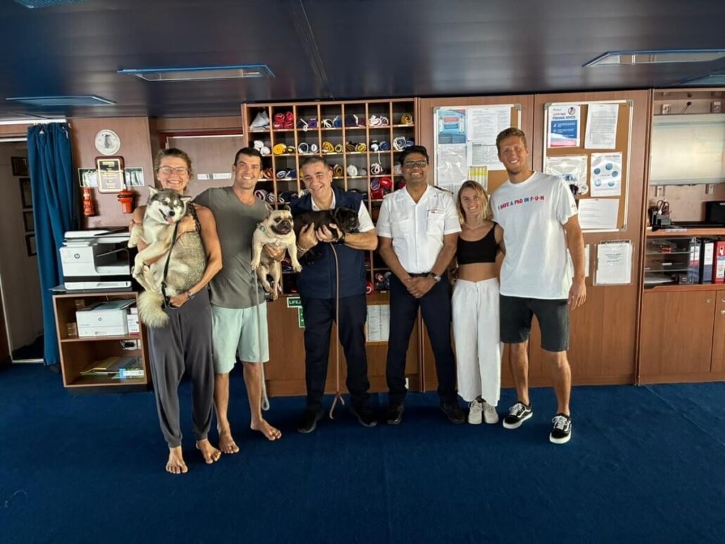 Six people stand in a row inside an office-like room; two people hold dogs rescued from a stranded catamaran. Shelves, organized files, and documents cover the wall behind them. Everyone is smiling and casually dressed except for two in uniforms.