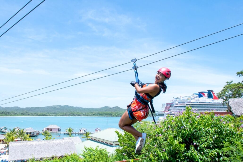 A person wearing a helmet and harness zip lines over a lush, green area with water and a large Carnival Cruise Line ship visible in the background under a blue sky, enjoying one of the thrilling shore excursions.