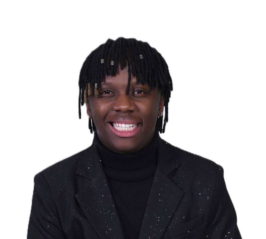 A person with medium-length black dreadlocks, wearing a black turtleneck and a black blazer with white speckles, smiles at the camera against a plain white background, ready to discuss the 2025 Carnival Cruise Director Schedule on ship.