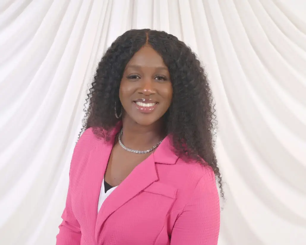 A woman with long, curly hair wears a pink blazer over a white top, a necklace, and hoop earrings. She is smiling and standing in front of a white, draped fabric background, ready to announce the Carnival Cruise Director Schedule.