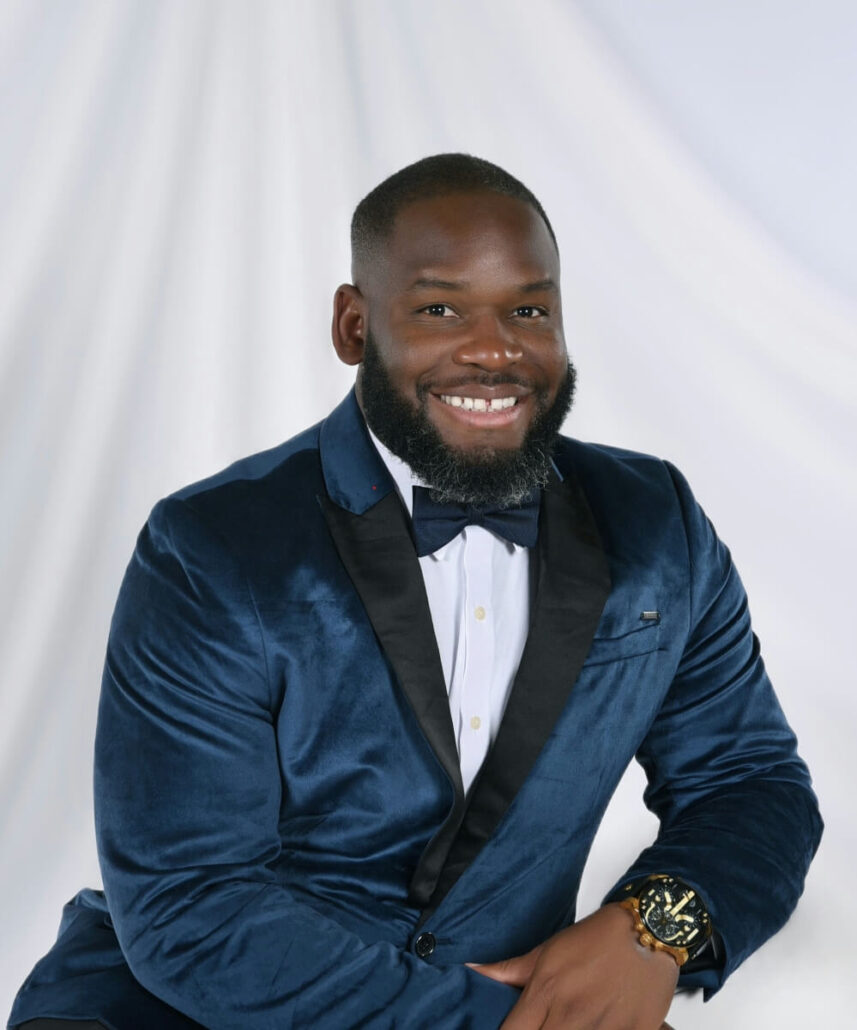 A man with a beard, possibly a Cruise Director, is sitting and smiling at the camera in a blue velvet suit jacket, white shirt, black bow tie, and gold wristwatch. The plain white background with draped fabric hints at a 2025 Carnival Cruise Director Schedule photo shoot.