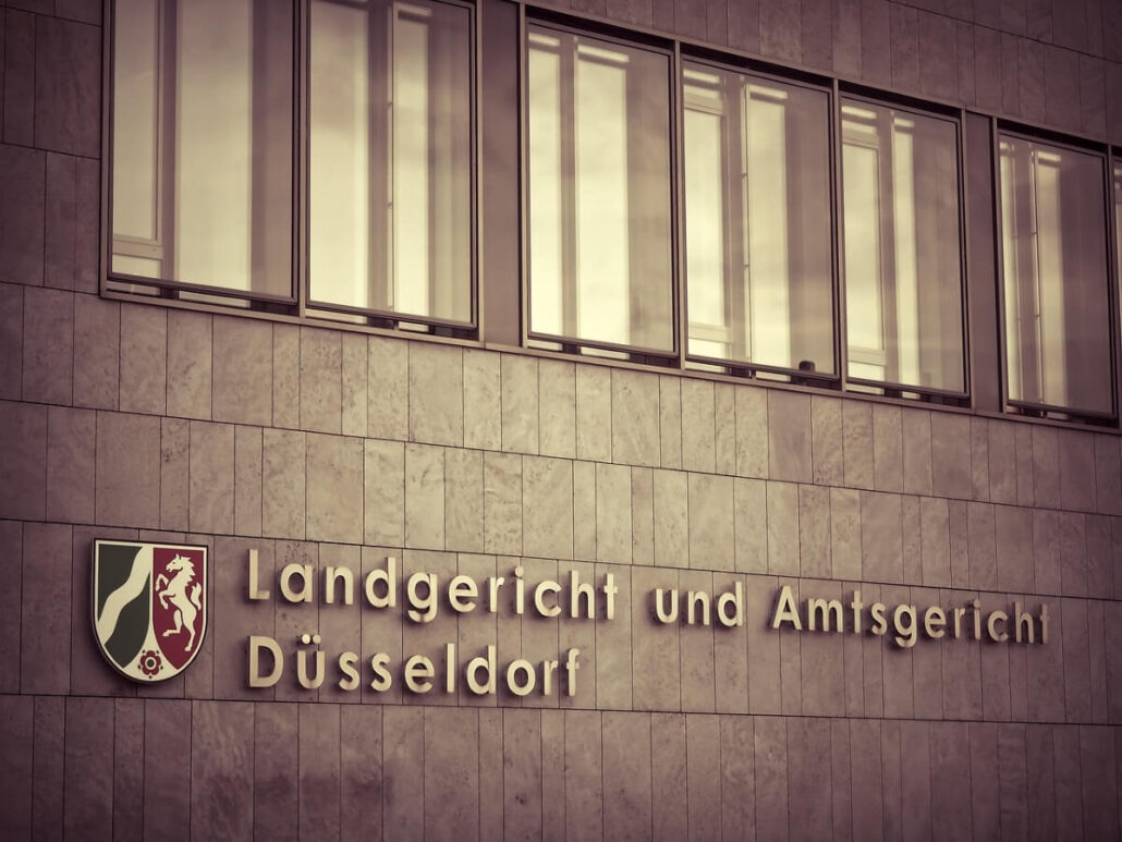 The exterior of a building with a sign reading "Landgericht und Amtsgericht Düsseldorf" next to a shield emblem, indicating it is a courthouse in Düsseldorf, Germany, possibly the site of an important cruise line court case. Several large windows sit above the sign.