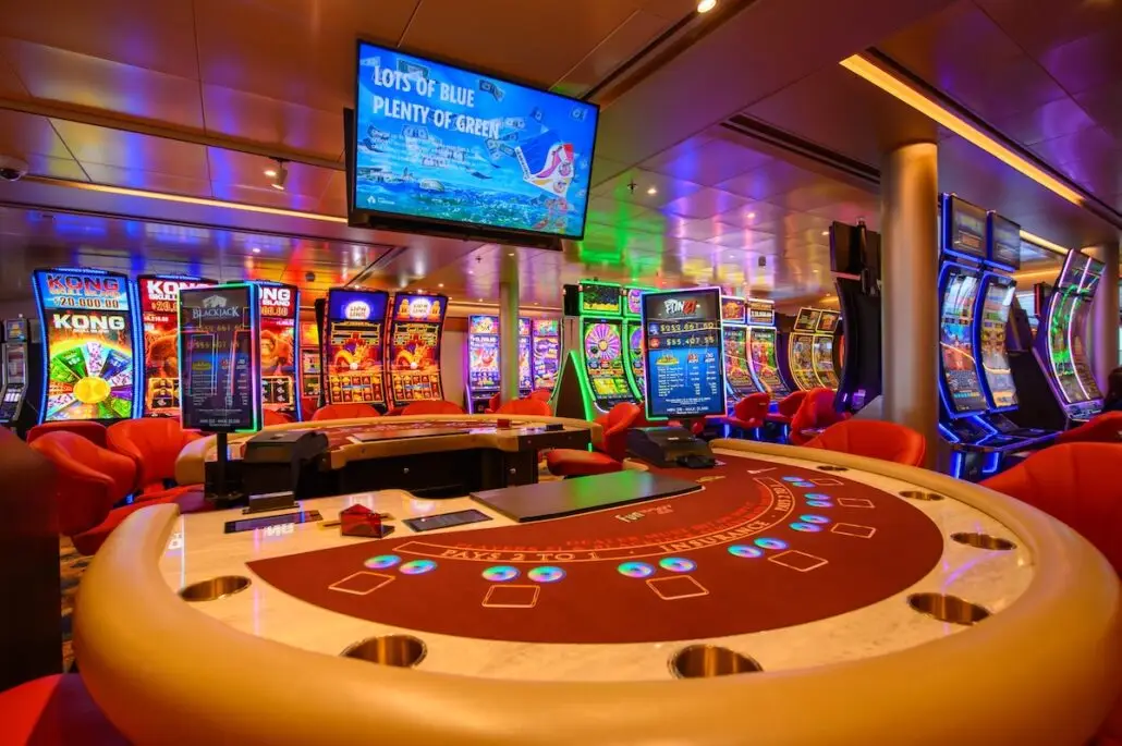 A brightly lit casino aboard Carnival Miracle features a round blackjack table in the foreground and rows of colorful slot machines behind. An overhead screen displays a game promotion, showcasing new features after the ship's recent relaunch.