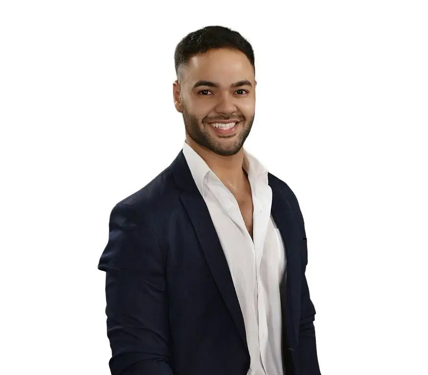 A man with short dark hair and a trimmed beard is smiling at the camera. He is wearing a white dress shirt under a dark blazer, resembling a professional cruise director from the 2025 Carnival Cruise Director Schedule. The background is plain white.