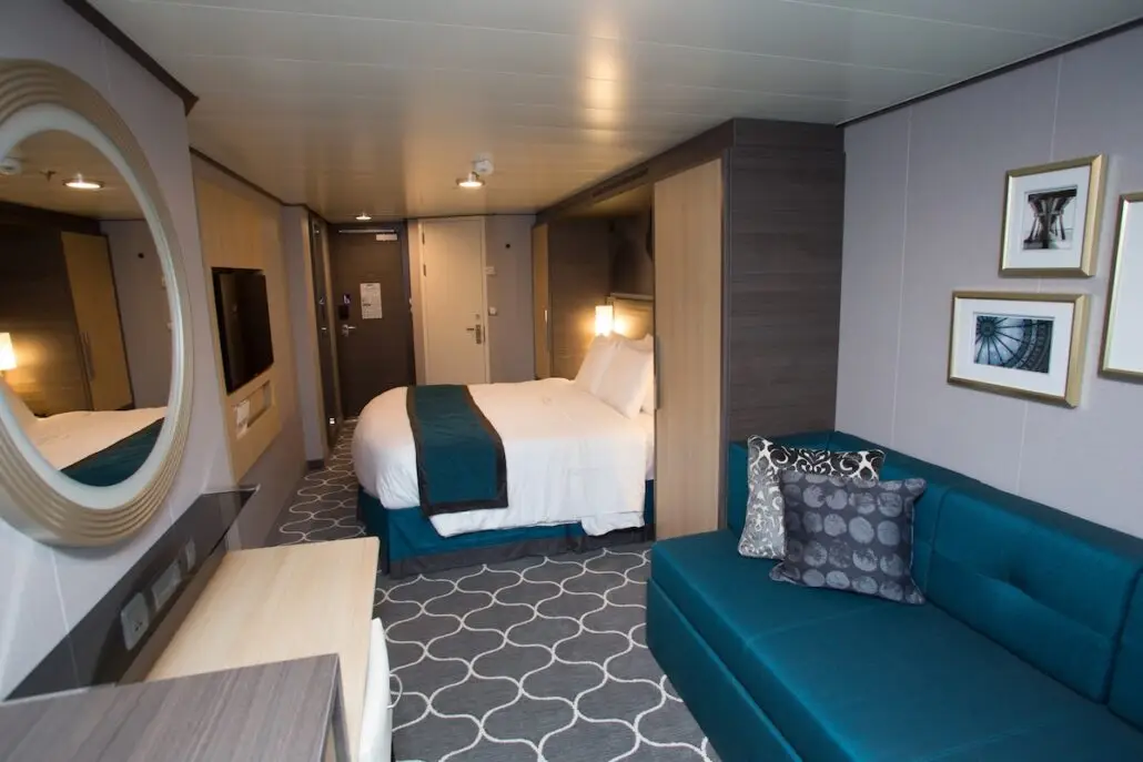 A modern cruise ship cabin, reminiscent of Royal Caribbean Oasis Class style, with a double bed, teal accents, wall-mounted TV, round mirror, desk, teal sofa, patterned carpet, and framed pictures on the wall—perfect for any cruise comparison.