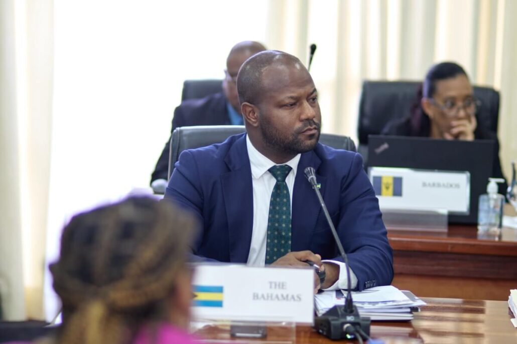 A man in a suit sits at a conference table with a microphone, labeled "The Bahamas." Papers and a nameplate are in front of him. Other people are seated in the background, one at a laptop labeled "Barbados," discussing Labor Law Violations.