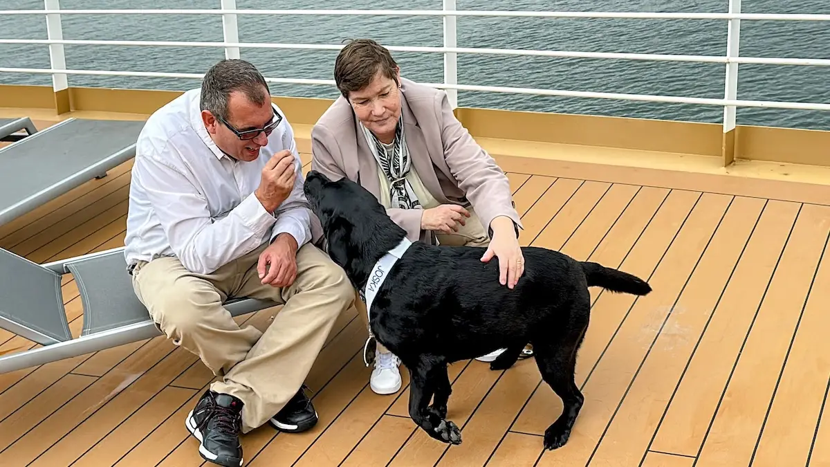 Two cruise guests relax on a Holland America deck, one holding something near their face while a black service dog in a harness stands nearby. Lounge chairs and a railing overlook the sparkling water.