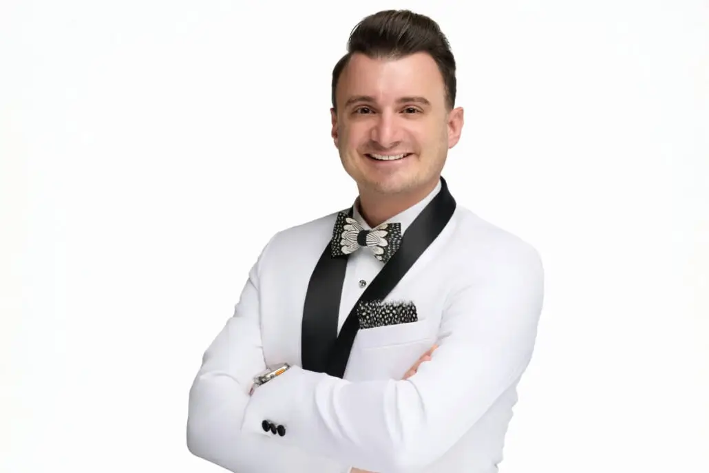 A man with neatly styled brown hair wears a white tuxedo with black lapels, a patterned bow tie, and a matching pocket square. He stands against a plain white background, smiling confidently like a 2025 Carnival Cruise Director.